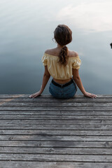 Fototapeta na wymiar Beautiful young woman in fashion blue jeans sits on a wooden pier near the lake