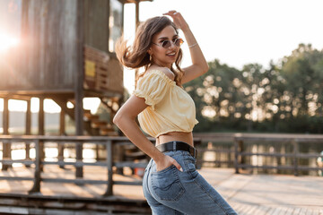 Beautiful hipster curly woman model with fashion sunglasses in blue jeans and fashionable summer top blouse walks in nature by the lake at sunset