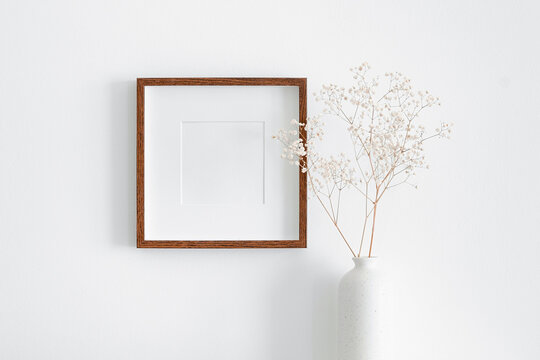 Square wooden frame with passepartout on white wall with dry gypsophila plant in vase. Blank mockup for artwork, print or photo presentation.