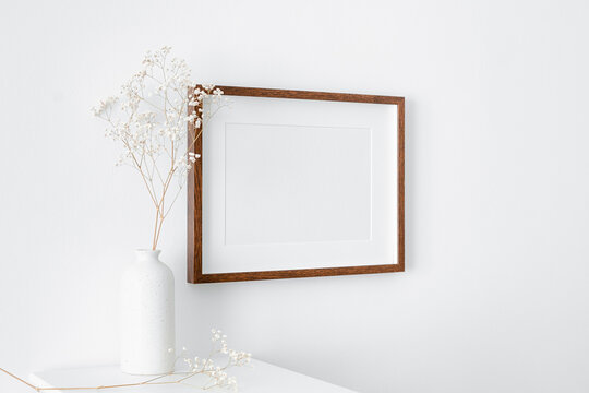 Blank landscape frame mockup on white wall with furniture and dry gypsophila plant decorations. Minimalistic interior with artwork mockup.