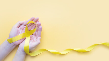 Hand holding yellow gold ribbon awareness on yellow background. Symbol for support suicide prevention, endometriosis, sarcoma bone cancer, bladder cancer, liver cancer and childhood cancer concept.