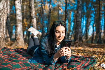 Portrait of a happy young woman reading a text message on a mobile phone, lying on a blanket in an autumn park. Communicate with friends and family using your smartphone. copy space