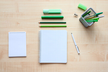 A squared notebook and green office supplies lie on a wooden desk. View from above. Selective focusing at the center of the frame.