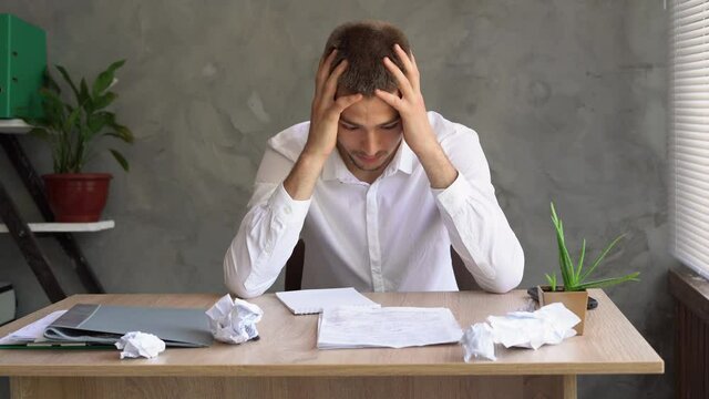 Portrait of an unmotivated businessman with a headache sitting in a company office. Solving financial problems. Creative crisis and depression. Employee burnout without inspiration.