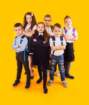 Group Of Happy Kids In Uniform With School Bags Hugging And Looking To Camera Over Yellow Background