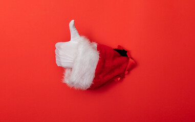 Santa Claus hand thumbs up through a hole in red paper background