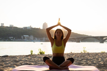 Woman in sport wear at sunset on beach sit on mat does yoga poses face to camera, calm and peaceful, positive emotions