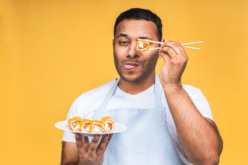 Young african american indian black man eating sushi using chopsticks over isolated yellow background. Cook preparing sushi.