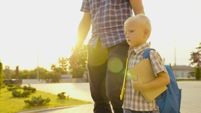Happy family, father, child go to school together. Dad is taking his little son with textbook to school. Schoolboy boy walking down street with backpack, holding his dad by hand. Preschool education.