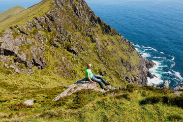 Bald male tourist in green shirt sitting on the edge of a rock. Achill island, county Mayo,...