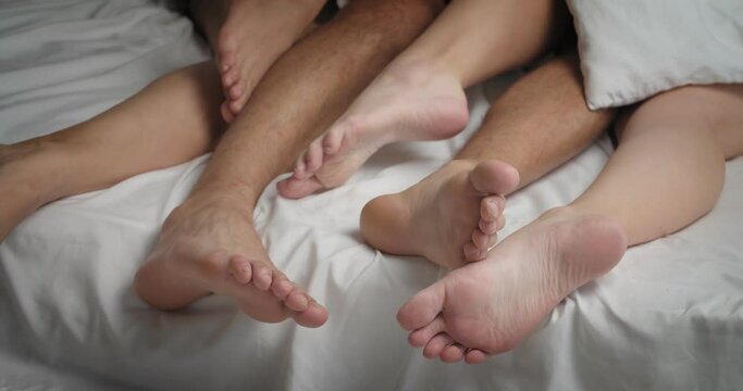 Feet of young man and two women lying in bed under blanket. Polyamory concept