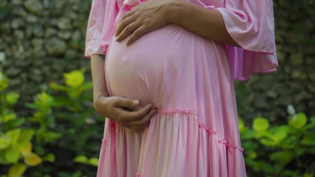 European pregnant girl in a pink dress gently touches her big stomach with her hands, stands on the back green garden