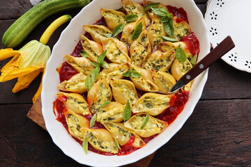 Big shell shape pasta stuffed with creamy soft cheese, zucchini and spinach sprinkled with parmesan cheese in baking dish wooden table, healthy dinner recipe, view flat lay