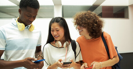  Three teenage multi-ethnic students chat on cell phones and talk and laugh with each other in the corridors of the school.