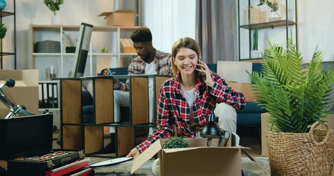 Likable confident young black-skinned guy measuring sizes of rack with tape measure when his cheerful pretty girlfriend unpacking cartons and talking on mobile,relocation concept