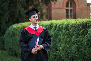 Portrait of handsome caucasian male graduate in graduation robe looking at campus
