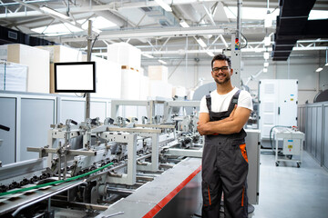 Portrait of an experienced worker in working uniform standing next to modern packaging machine in printing factory.