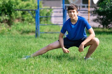 teenage boy exercising outdoors, sports ground in the yard, he squats and does a warm-up, healthy lifestyle