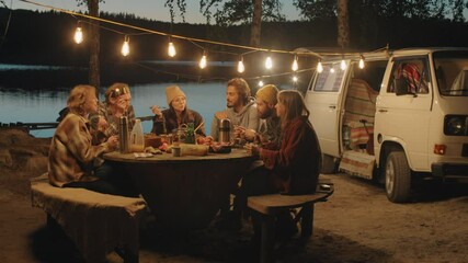 Group of young men and women sitting together at table under garland, eating meal and having discussion at late dinner on campsite