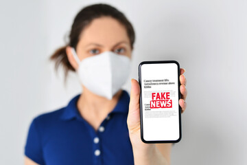 Young woman wearing medical mask and holding a smartphone with fake news in front. Propaganda,...