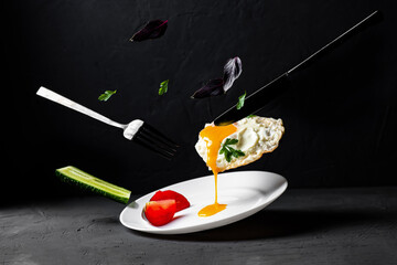 Fried egg levitation. Dark food photo. A chicken egg with liquid yolk, leaves of basil, dill, cucumber falls on a white plate. Breakfast creative concept.