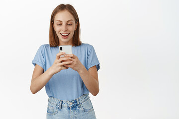 Excited smiling blond girl taking photo on mobile phone. Young happy woman looking amazed at smartphone screen, found smth at online store, standing against white background