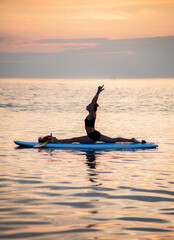 a female makes yoga asana on the SUP board at the orange light of sunrise