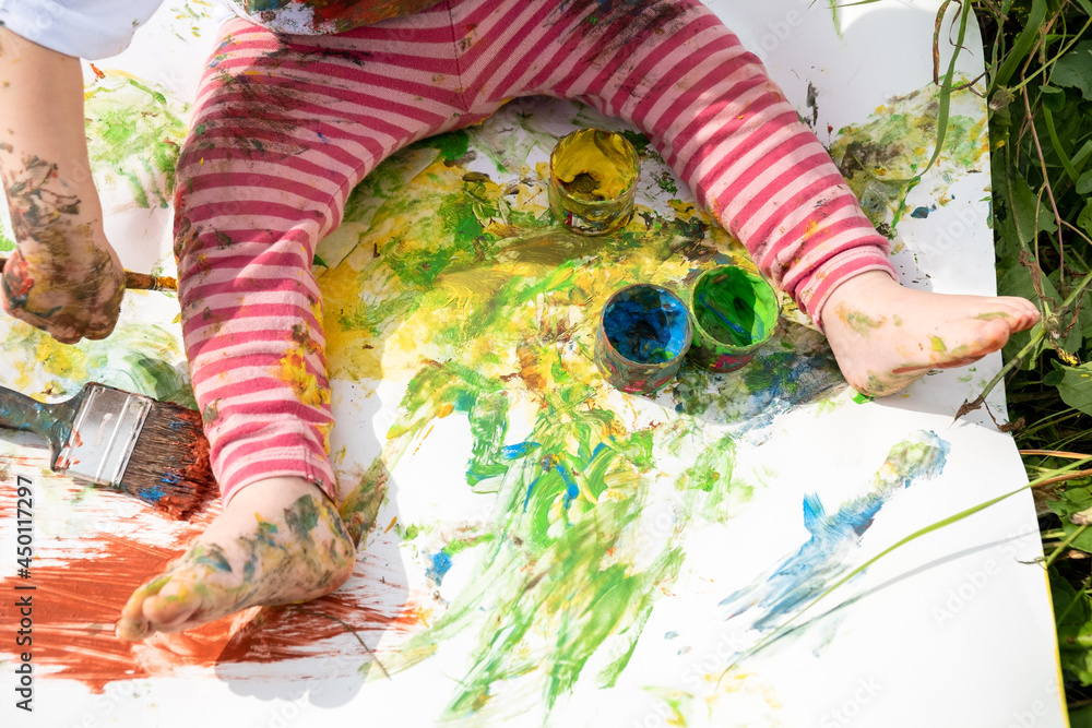 Wall mural Baby draws with paints sitting on a white sheet of paper, top view.