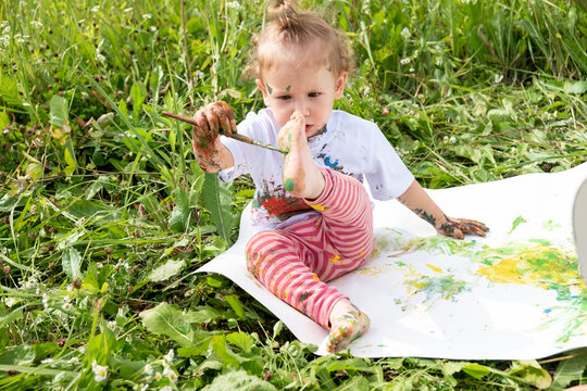 A small child draws with paint on his foot, sitting on a sheet of white paper.
