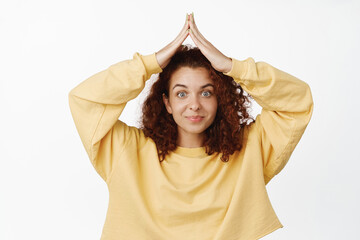 Cute and silly redhead woman making small house rooftop gesture above head, hand roof on top and smiling, standing over white background