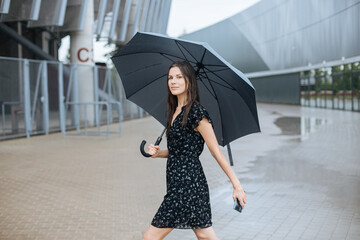 Woman walking with black umbrella. Urban city lifestyle. A female in a dress. Fall shopping holidays. Attractive businesswoman outdoor fashion. City life during rain.