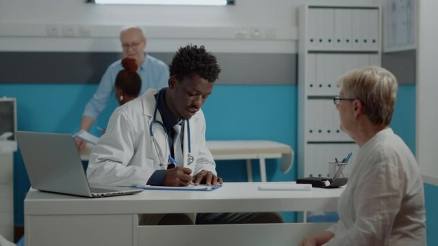 African american doctor writing checkup on notebook while consulting senior woman at desk. Black medic checking on patient healthcare while nurse and elder man talking in background
