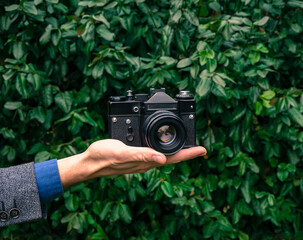 Male hand holding a vintage film camera in the air. Green nature in the background.