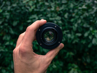 Male hand holding a vintage camera lens in the air. Green leaves in the background.