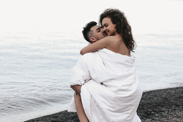 A couple in love are walking together hugging spend the night covered with a white blanket on the ocean beach on a romantic honeymoon in nature. Passionate man and woman of mixed diverse races