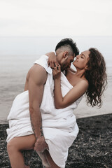 Obraz premium A couple in love are walking together hugging spend the night covered with a white blanket on the ocean beach on a romantic honeymoon in nature. Passionate man and woman of mixed diverse races