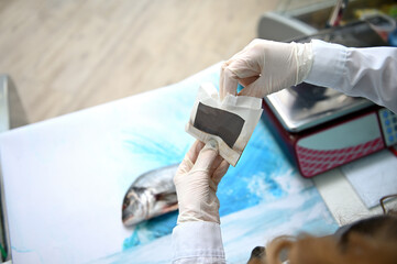 Focus on the chef's gloved hands. The fishmonger holds a paper bag of spices and sprinkles the seasoning on the dorado fish on the tabletop.