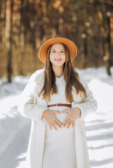 Beautiful happy pregnant woman in hat and sweater standing in the field and holding her belly at winter time