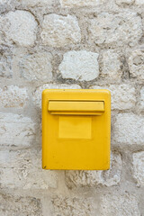 A yellow mailbox on a light stone wall. Box for letters and postcards.