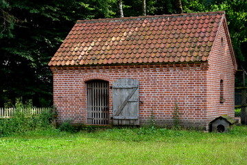 Fototapeta na wymiar A close up on a barn, pen, or shelter made out of red bricks, tiles, and wooden elements standing in the middle of a well maintained lawn with a small dog house to the side seen on a sunny summer day
