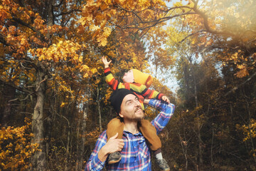 Cute little child boy sitting on his father's shoulders on background of golden autumn trees in...
