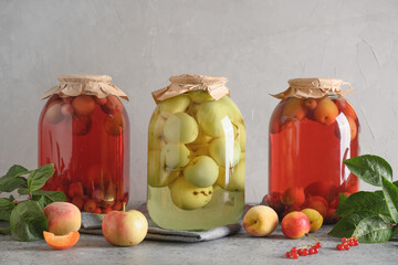 Three homemade canned fruits apple and cherry compote in large glass jars on gray table. - obrazy, fototapety, plakaty