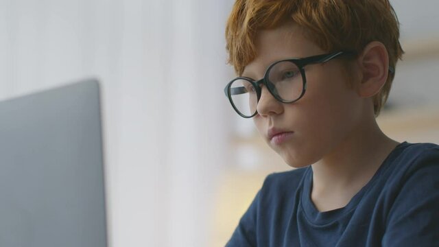 Close up portrait of focused little redhead boy typing on laptop, studying online distantly from home