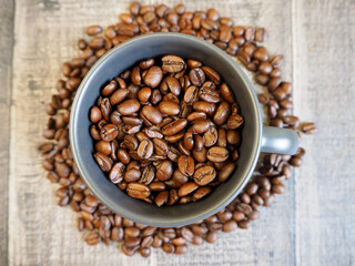 a dark gray mug filled with roasted whole coffee beans stands on a wooden background with coffee beans at the bottom of the mug. top view