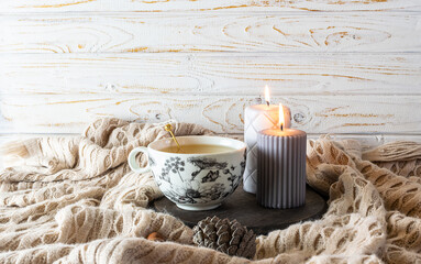  Creative hygge style composition with cup of herbal tea, candles and warm plaid on white wooden background. Cozy home interior.