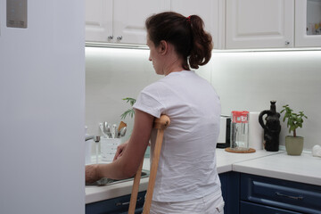 Woman on crutches washes the dishes in the kitchen. Temporary limitation of the physical capabilities of a person with a broken leg
