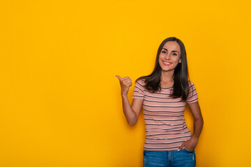 Cute excited smiling young brunette girl is pointing away for some advertisement while stands on yellow wall background