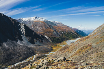 Picturesque highland landscape with mountain river and lake in valley among high mountains with snow in autumn colors in sunshine. Awesome alpine view to motley mountain valley in sunlight in fall.