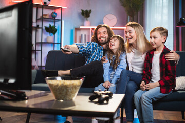 Cheerful man and woman with two children sitting in hugs on comfy sofa and using remote control...