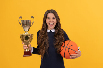 happy teen girl hold basketball ball and champion cup on yellow background, happiness